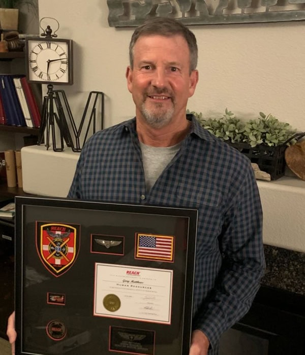Man holding framed military and achievement awards, including a fire department badge and American flag, in an office setting.