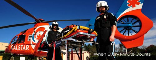Two emergency medical personnel preparing a Searle Starter helicopter for patient transport, with a bright orange helicopter featuring medical symbols and a clear blue sky background.