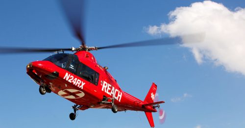 Red REACH Air Medical Searle Starter helicopter flying in clear blue sky, used for emergency medical transport under REACH Air Medical services.